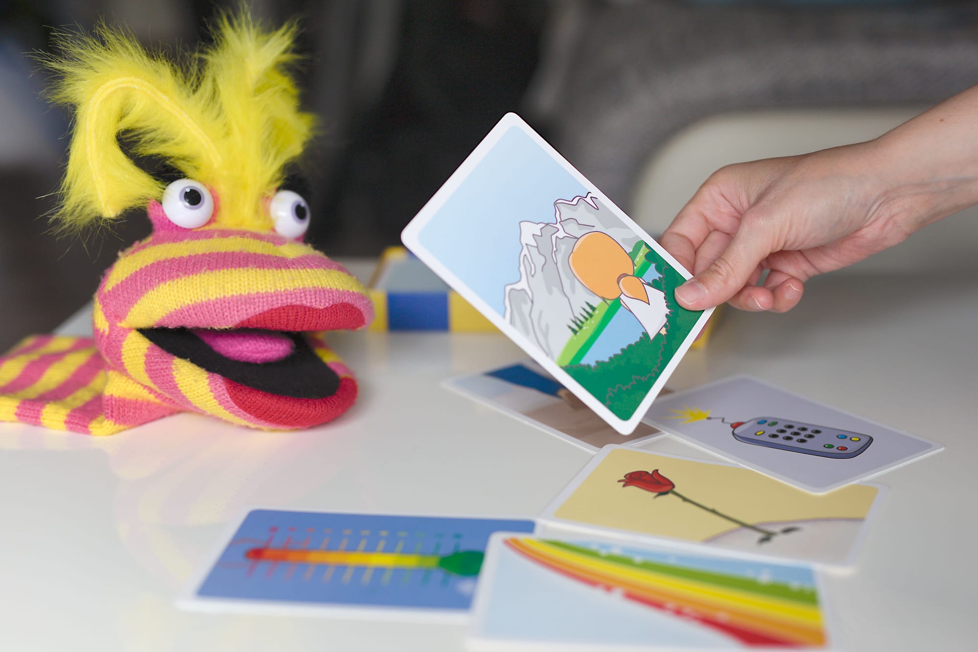 Therapiekarten liegen auf dem Tisch, Hand hebt eine Therapiekarte auf und eine Handpuppe liegt hinten auf dem Tisch.